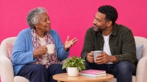 An older mother and her adult son share an attentive conversation against a bright pink background.