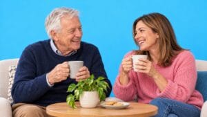 An older father and his adult daughter smile together over tea against a bright blue background.